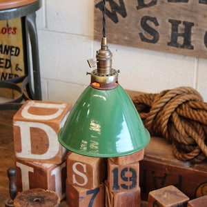 May include: A green enamel pendant light fixture with a conical shade and a metallic brass-toned socket assembly. The fixture features a black twisted fabric cord and a small turn-key switch located on the side of the brass hardware component.