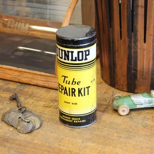 以下が含まれることがあります： 黄色いラベルと黒い文字が入った、ヴィンテージのDunlop Tube Repair Kitの缶。金属製の鍵と、木製の車輪が付いた小さな緑色の玩具車も隣にあります。木製の表面に置かれています。