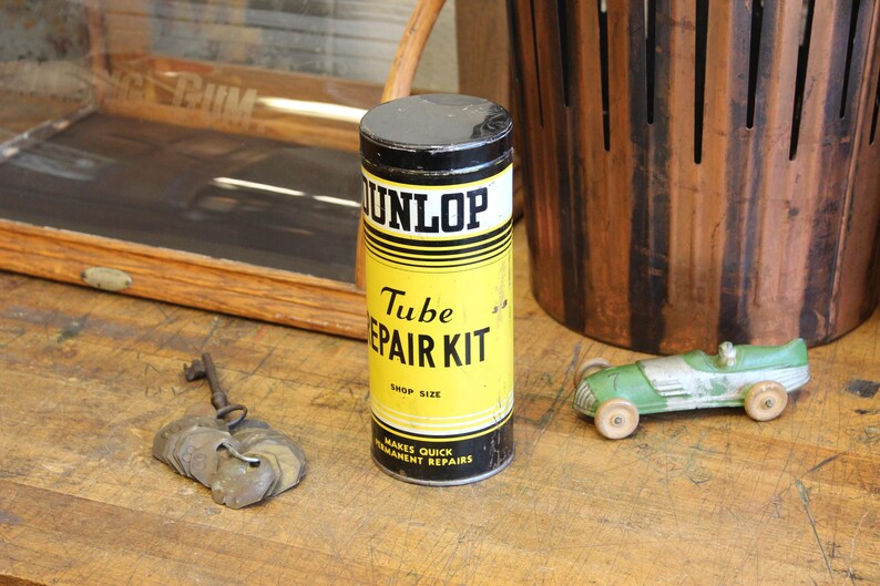 以下が含まれることがあります： 黄色いラベルと黒い文字が入った、ヴィンテージのDunlop Tube Repair Kitの缶。金属製の鍵と、木製の車輪が付いた小さな緑色の玩具車も隣にあります。木製の表面に置かれています。