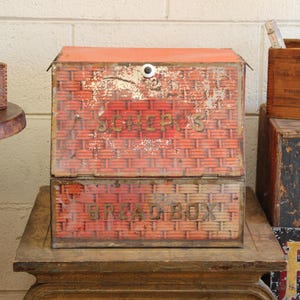 May include: Vintage red and orange metal bread box with a woven basket design. The box has the word "SCHEPPS" on the top and "BREAD BOX" on the bottom. The bread box is sitting on a wooden table.