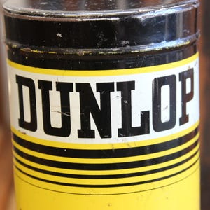 以下が含まれることがあります： 黒い蓋と黄色の本体が付いた円筒形の容器。 「DUNLOP」の文字が、白い長方形のラベルに太字の黒い文字で印刷されています。 容器には黒と黄色の水平ストライプがあります。