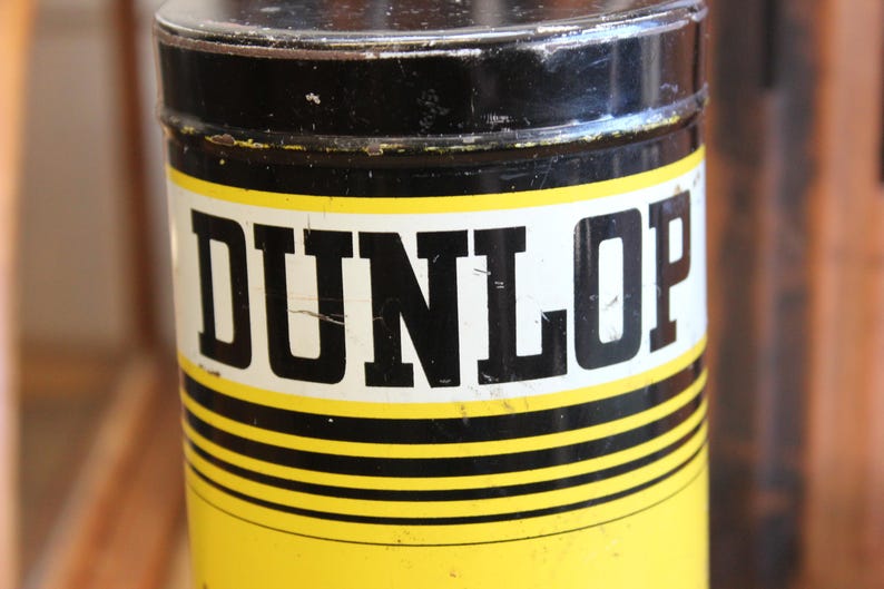 以下が含まれることがあります： 黒い蓋と黄色の本体が付いた円筒形の容器。 「DUNLOP」の文字が、白い長方形のラベルに太字の黒い文字で印刷されています。 容器には黒と黄色の水平ストライプがあります。