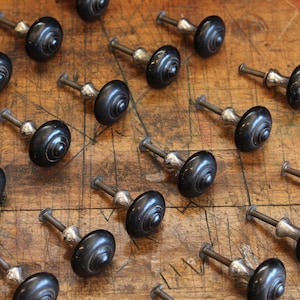 May include: A collection of round black cabinet knobs with silver-toned metal bases and threaded mounting screws. Each knob features a smooth, circular top with a central raised point, arranged in a grid pattern across the frame.