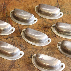 May include: A collection of antique, silver-toned metal drawer pulls. Each pull is a half-moon shape with a white rim and two screw holes. The pulls are arranged on a wooden surface.