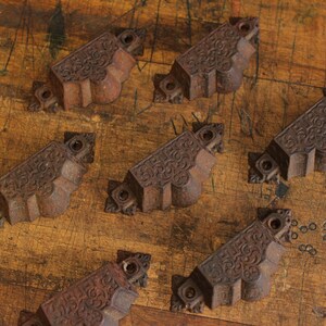 May include: A collection of six antique, cast iron drawer pulls. Each pull is a dark, rusty brown color with ornate, scrolled detailing. The pulls are rectangular with decorative end pieces and screw holes for mounting.