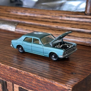 May include: A teal-colored die-cast model car with its hood open, revealing engine details. The classic car has black wheels and sits on a wooden surface. The car's design is reminiscent of vintage automobiles.
