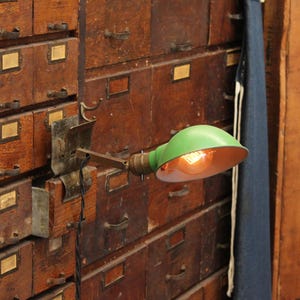 May include: A green metal wall-mounted lamp with a dome-shaped shade and a white interior. The lamp features a metallic arm attached to a rectangular mounting plate and includes a glowing light bulb. A black twisted cord hangs from the fixture.