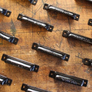 May include: A collection of black metal drawer pulls with the word "HAMILTON" embossed on each. The pulls are rectangular with rounded edges and two screw holes on each end. They are arranged on a weathered wooden surface.