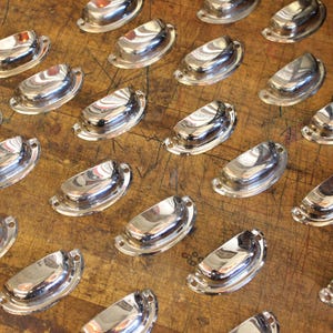May include: A collection of shiny, silver-colored cup pulls arranged on a wooden surface. Each pull has a curved, half-moon shape with two small holes for mounting. The pulls reflect the light, creating a bright and polished appearance.