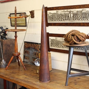 May include: A tall, dark brown wooden cone stands on a wooden table. A wooden tripod stand with a round top and a wooden spool holder are to the left. A coil of rope sits on a small stool to the right.