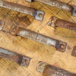 May include: A collection of six rusty metal drawer pulls with the word "HAMILTON" embossed on each. The pulls are arranged in a staggered pattern on a wooden surface.
