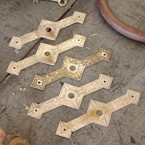 May include: Four antique silver-toned metal drawer pulls with an intricate diamond-shaped design and a circular hole in the center. The pulls are arranged in a staggered pattern on a wooden surface.