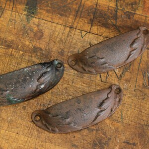 May include: Three antique, cast iron drawer pulls with a weathered, distressed finish. The pulls are a dark, mottled brown with hints of green and white, and feature a curved, scoop-like design. They are set against a worn, wooden surface.