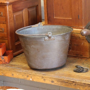 May include: A vintage copper bucket with a metal handle. The bucket is sitting on a wooden surface.