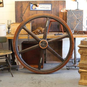 May include: Large, antique wooden wheel with eight spokes and a dark brown finish. The wheel has a lighter brown rim and a central hub with a copper-colored ring. The wheel is displayed indoors, leaning against a wall.