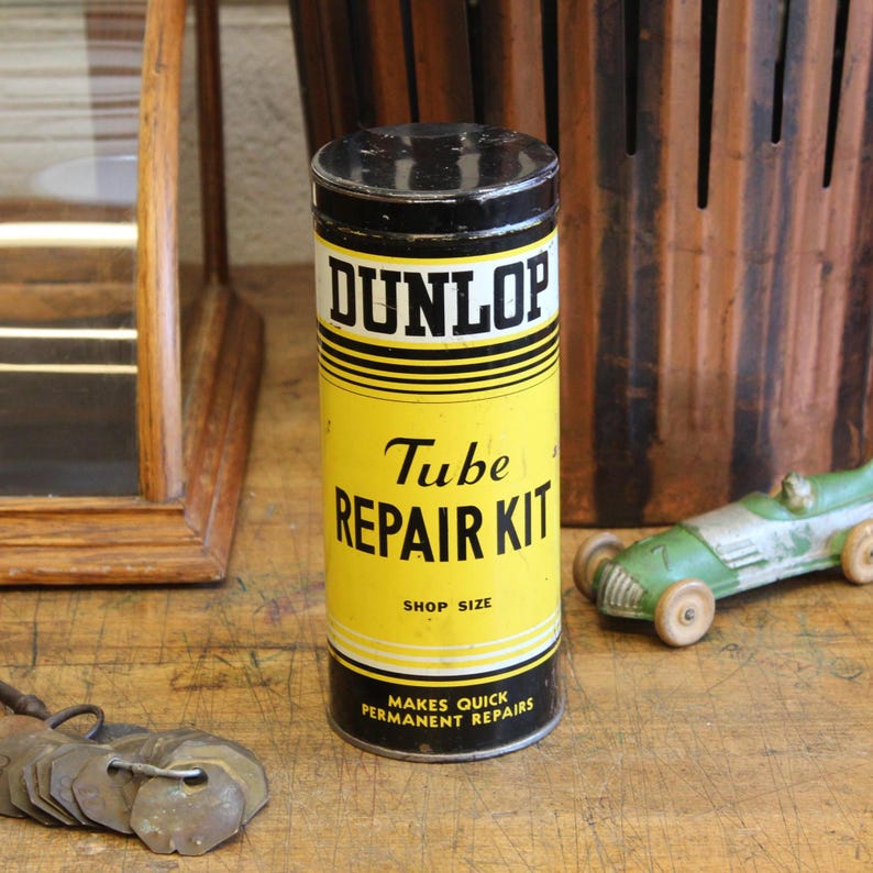 以下が含まれることがあります： 黒い蓋とベース、黄色のボディのヴィンテージDunlop Tube Repair Kit缶。上部に「DUNLOP」、下に「Tube REPAIR KIT」の文字。缶には「SHOP SIZE」と「MAKES QUICK PERMANENT REPAIRS」と書かれています。