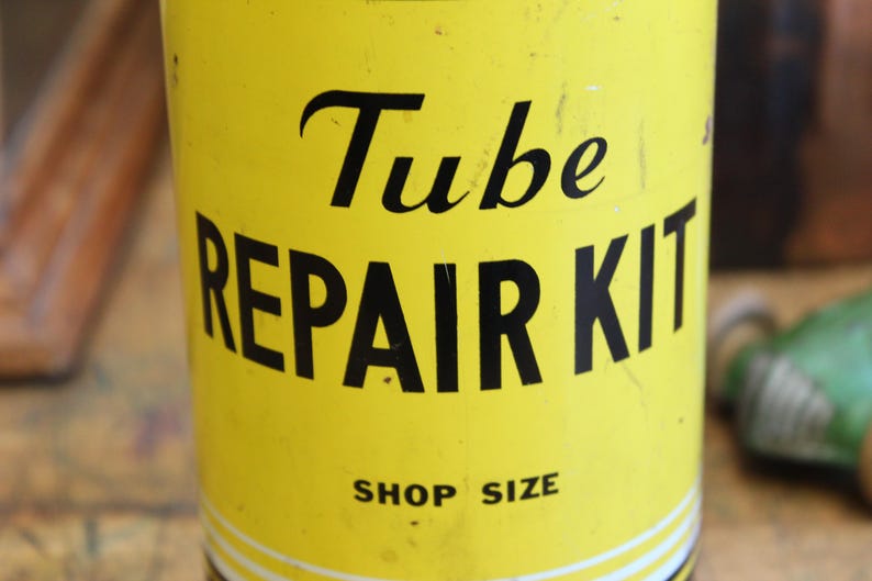 以下が含まれることがあります： 黒い文字で「Tube REPAIR KIT」と書かれた黄色のチューブ修理キット。「SHOP SIZE」の文字も含まれています。少し使い込まれたような、ヴィンテージ感のある外観です。