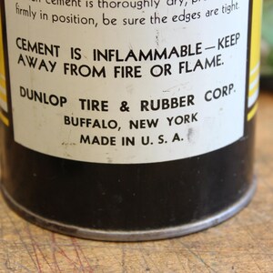 以下が含まれることがあります： ヴィンテージのDunlop Tire & Rubber Corp.のセメント缶のクローズアップ。ラベルには「CEMENT IS INFLAMMABLE - KEEP AWAY FROM FIRE OR FLAME」と書かれています。缶は黒色で、黄色と白のラベルが付いており、「MADE IN U.S.A.」と印刷されています。