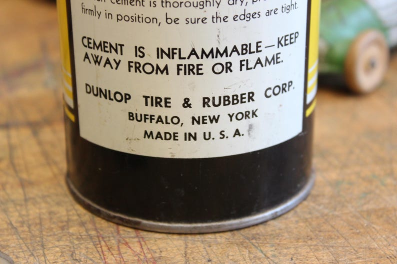 以下が含まれることがあります： ヴィンテージのDunlop Tire & Rubber Corp.のセメント缶のクローズアップ。ラベルには「CEMENT IS INFLAMMABLE - KEEP AWAY FROM FIRE OR FLAME」と書かれています。缶は黒色で、黄色と白のラベルが付いており、「MADE IN U.S.A.」と印刷されています。
