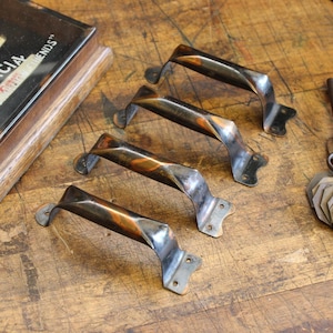 May include: Four antique metal door handles with a twisted design. The handles have a dark finish with hints of copper and are mounted on a wooden surface. Each handle has a curved grip and mounting plates with screw holes.