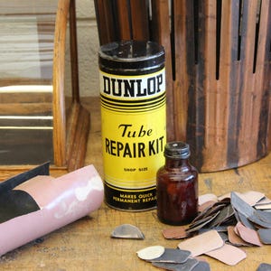 以下が含まれることがあります： 黄色と黒のラベルが付いたヴィンテージのダンロップチューブ修理キット、小さな茶色のボトル、さまざまなパッチ。 このキットはショップサイズで、迅速かつ恒久的な修理のために設計されています。 木の表面にアイテムがあります。