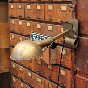 May include: A brass clamp-on lamp with a dome-shaped shade and an adjustable arm. The lamp features a metallic finish with visible signs of wear and a black fabric-covered electrical cord extending downward from the base of the lamp head.