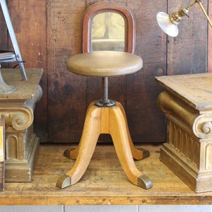 May include: An antique wooden adjustable stool with a round, tan seat and a small, arched mirror back. The stool has a light brown wooden base with brass accents on the feet. The metal screw mechanism allows for height adjustment.