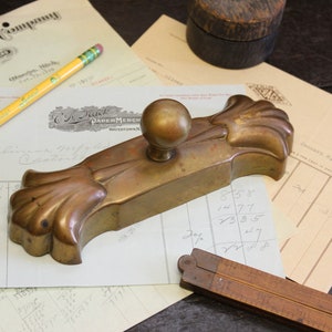 May include: A brass inkwell with a decorative floral design and a round knob on top. The inkwell is sitting on a piece of paper with handwritten numbers and a ruler.