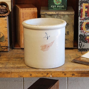 Puede incluir: Un tarro de gres blanco roto vintage con un borde redondeado. Presenta un número 4 azul y un diseño de ala marrón rojizo. El tarro cilíndrico se encuentra sobre una superficie de madera.