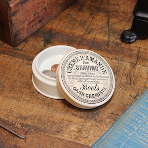 May include: An open, off-white ceramic shaving cream container with a lid. The lid features the text "Creme D'Amande for Shaving" and "Boots Cash Chemists". The container holds several coins. The background is a wooden surface.