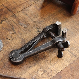 May include: An antique, black metal clamp tool with a screw mechanism and a handle. The tool has a weathered appearance, suggesting age and use. The clamp is designed to hold objects securely. The tool is on a wooden surface.