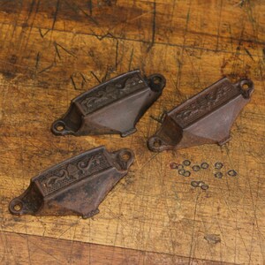 May include: Three antique cast iron drawer pulls with decorative scrollwork and mounting holes. The pulls are dark brown with a weathered appearance, and are arranged on a wooden surface with small washers.