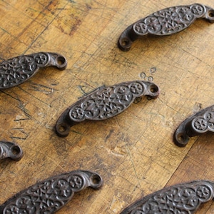 May include: Close-up of several antique-style, cast iron drawer pulls. The pulls are dark brown with an ornate, floral design. They are arranged on a light brown, wooden surface with visible grain and scratches.
