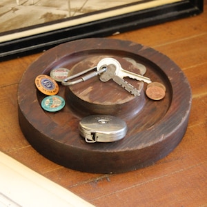 May include: A round, dark brown wooden tray with a central raised area. The tray holds keys, coins, and several colorful enamel pins. A small, silver tape measure is also present, suggesting a nautical or vintage theme.