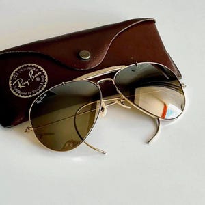 K&ouml;nnte beinhalten: Goldfarbene Aviator-Sonnenbrille mit braunen Gl&auml;sern liegt auf einem braunen Lederetui. Das Etui hat einen Druckknopfverschluss und ein Ray-Ban-Logo. Die Sonnenbrille hat ein klassisches Design.