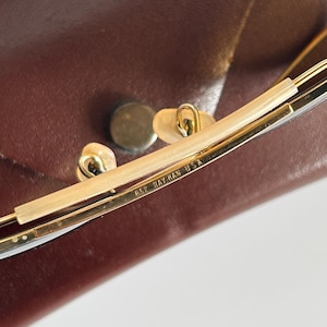 K&ouml;nnte beinhalten: Nahaufnahme einer Ray-Ban USA Brille. Die Brille hat einen goldfarbenen Rahmen mit einem hellfarbigen Steg und Nasenpads. Die Brille liegt auf einem braunen Lederetui.