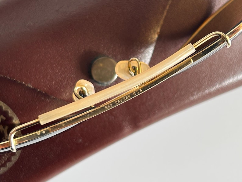 K&ouml;nnte beinhalten: Nahaufnahme einer Ray-Ban USA Brille. Die Brille hat einen goldfarbenen Rahmen mit einem hellfarbigen Steg und Nasenpads. Die Brille liegt auf einem braunen Lederetui.
