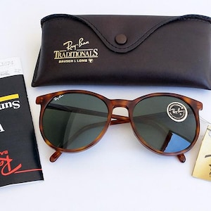 Puede incluir: Gafas de sol Ray-Ban con montura carey y lentes oscuras, acompa&ntilde;adas de un estuche de cuero y documentaci&oacute;n. Las gafas tienen una montura redonda y una pegatina en la lente. El estuche es de color marr&oacute;n oscuro.