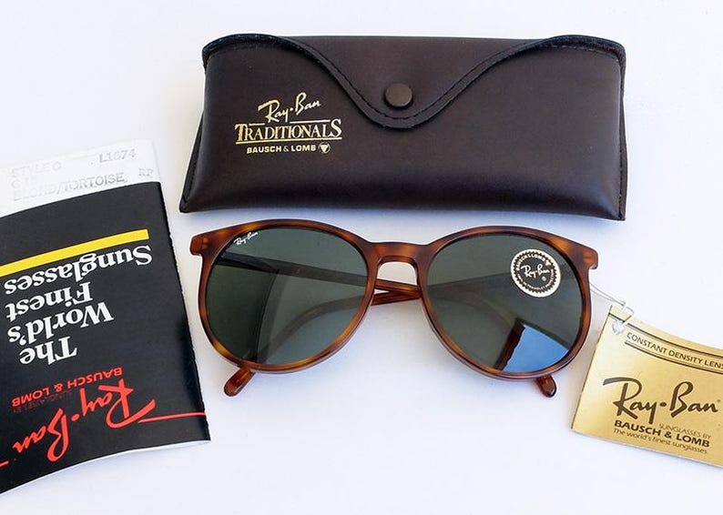 Puede incluir: Gafas de sol Ray-Ban con montura carey y lentes oscuras, acompa&ntilde;adas de un estuche de cuero y documentaci&oacute;n. Las gafas tienen una montura redonda y una pegatina en la lente. El estuche es de color marr&oacute;n oscuro.
