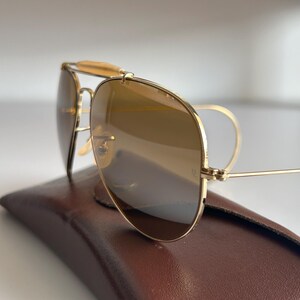 K&ouml;nnte beinhalten: Goldfarbene Aviator-Sonnenbrille mit braun get&ouml;nten Gl&auml;sern liegt auf einem braunen Lederetui. Die Sonnenbrille hat einen Doppelsteg und einen Stirnb&uuml;gel. Die Gl&auml;ser reflektieren die Umgebung.