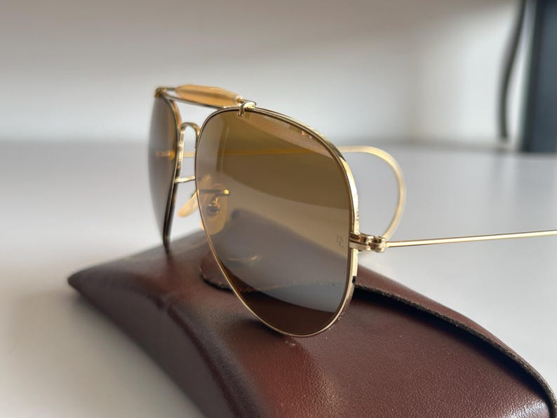 K&ouml;nnte beinhalten: Goldfarbene Aviator-Sonnenbrille mit braun get&ouml;nten Gl&auml;sern liegt auf einem braunen Lederetui. Die Sonnenbrille hat einen Doppelsteg und einen Stirnb&uuml;gel. Die Gl&auml;ser reflektieren die Umgebung.