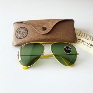 Puede incluir: Gafas de sol aviador amarillas con lentes verdes y una funda de cuero marr&oacute;n. Las gafas de sol tienen el logotipo de Ray-Ban en las lentes y la funda tiene el logotipo de Ray-Ban en la parte delantera.