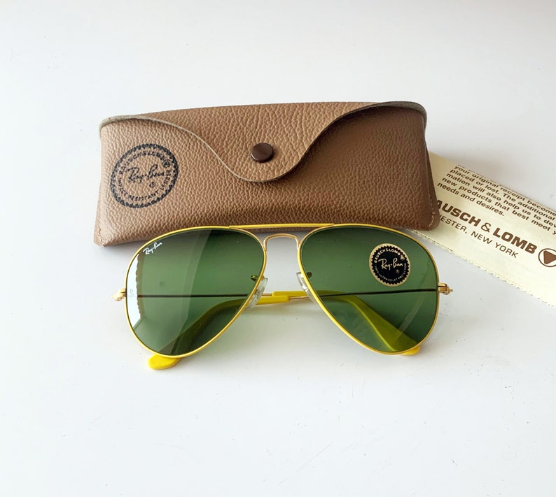 Puede incluir: Gafas de sol aviador amarillas con lentes verdes y una funda de cuero marr&oacute;n. Las gafas de sol tienen el logotipo de Ray-Ban en las lentes y la funda tiene el logotipo de Ray-Ban en la parte delantera.