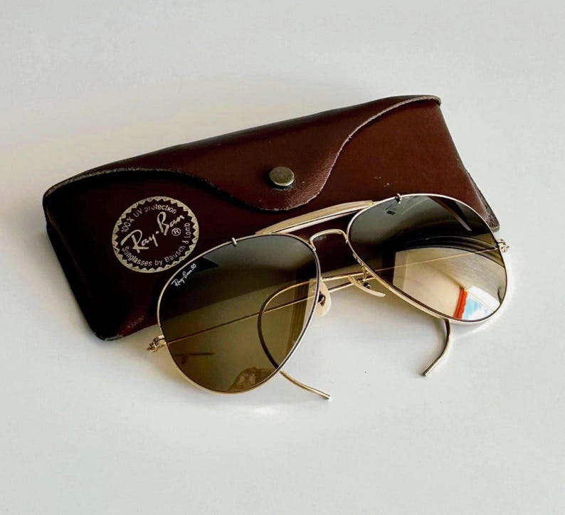 K&ouml;nnte beinhalten: Goldfarbene Aviator-Sonnenbrille mit braunen Gl&auml;sern liegt auf einem braunen Lederetui. Das Etui hat einen Druckknopfverschluss und ein Ray-Ban-Logo. Die Sonnenbrille hat ein klassisches Design.
