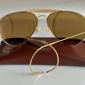K&ouml;nnte beinhalten: Goldfarbene Aviator-Sonnenbrille mit braunen Gl&auml;sern liegt auf einem braunen Lederetui. Die Sonnenbrille hat einen Doppelsteg und einen hellfarbigen B&uuml;gel &uuml;ber der Nase. Die Gl&auml;ser sind leicht gew&ouml;lbt.