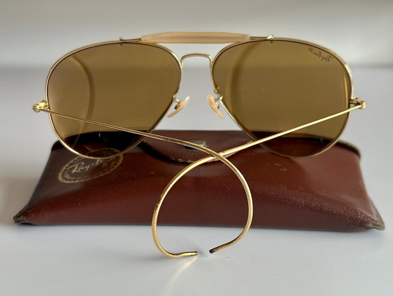 K&ouml;nnte beinhalten: Goldfarbene Aviator-Sonnenbrille mit braunen Gl&auml;sern liegt auf einem braunen Lederetui. Die Sonnenbrille hat einen Doppelsteg und einen hellfarbigen B&uuml;gel &uuml;ber der Nase. Die Gl&auml;ser sind leicht gew&ouml;lbt.