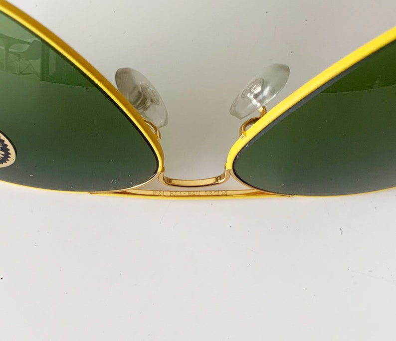 Puede incluir: Gafas de sol aviador amarillas y doradas con lentes verdes. Las gafas tienen el logotipo de Ray-Ban en el lateral.