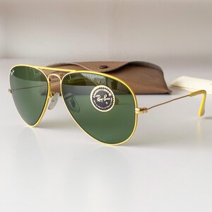 Puede incluir: Gafas de sol aviador amarillas con lentes verdes y un logotipo de Ray-Ban en la lente derecha.