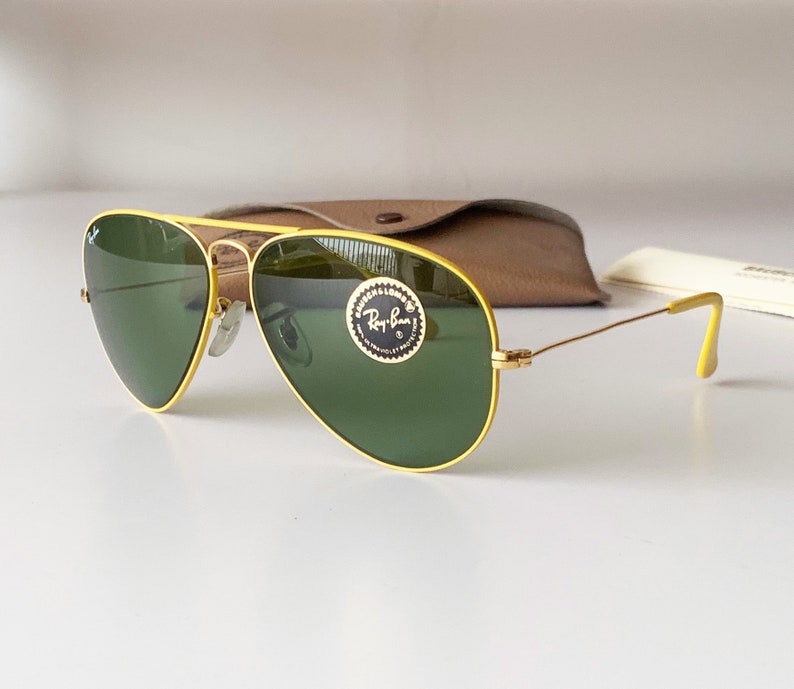 Puede incluir: Gafas de sol aviador amarillas con lentes verdes y un logotipo de Ray-Ban en la lente derecha.