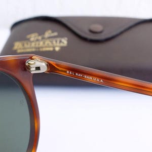 Puede incluir: Primer plano de gafas de sol Ray-Ban con montura carey y lentes verdes. La patilla derecha est&aacute; marcada con "B&L RAY-BAN U.S.A.". En el fondo, un estuche de cuero marr&oacute;n con letras doradas.